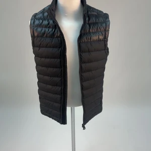 Svart dunväst från J Lindeberg  - Snygg svart dunväst från J Lindeberg med dragkedja framtill. Perfekt för lager-på-lager under kyliga dagar. Västen har en stilren design med quiltade paneler och hög krage.