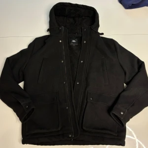 Fodrad Jacka - Säljer en svart fodrad jacka med huva och dragkedja. Jackan har långa ärmar och är fodrad med mjuk fleece för extra värme. Perfekt för kyliga dagar!