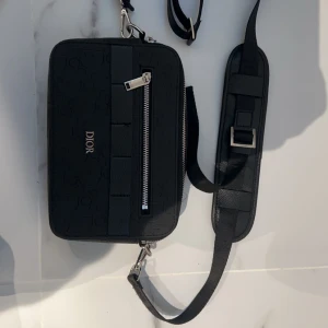 Svart axelväska från Dior - Snygg svart axelväska från Dior med ett stilrent mönster och en praktisk dragkedja framtill. Väskan har en justerbar axelrem och en elegant design som passar perfekt för alla tillfällen. Perfekt för dig som vill ha en lyxig touch i vardagen. Fler bilder privat 