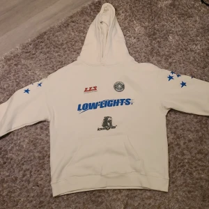 Vit hoodie från Lowlights studios - Säljer en vit hoodie från Lowlights med blå stjärnor på ärmarna och tryck på framsidan. Hoodien har en klassisk design med en stor ficka framtill och en bekväm huva. Perfekt för en avslappnad stil.