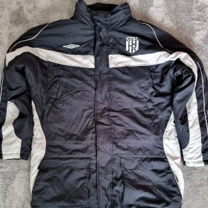 Svart och vit jacka från Umbro - Säljer en svart och vit jacka från Umbro med logotyp på bröstet och ett emblem. Jackan har långa ärmar och dragkedja framtill. Perfekt för sportiga aktiviteter eller som en casual jacka. Storlek XL.