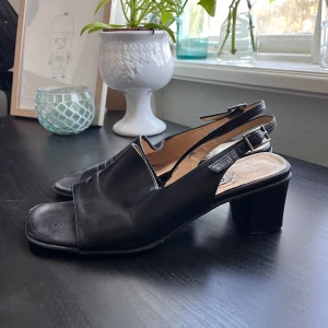 Svarta sandaler med klack från Giko - Stilrena svarta sandaler från Giko med öppen tå och justerbar rem runt hälen. Skorna har en bekväm blockklack och är tillverkade i skinn, vilket ger en elegant look. Perfekta för varmare dagar.