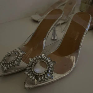 Silvriga klackskor med dekorativ detalj - Eleganta silvriga klackskor med en glittrig dekorativ detalj framtill. Skorna har en spetsig tå och transparenta sidor som ger en modern touch. Perfekta för speciella tillfällen.
