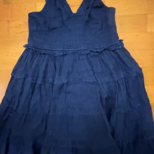 Mörkblå klänning med volangdetaljer - Säljer en mörkblå klänning med volangdetaljer och smockad överdel. Klänningen har breda axelband och en luftig kjol som ger en avslappnad look. Perfekt för sommarens varma dagar!