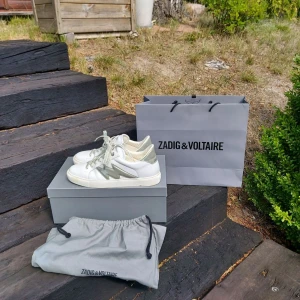 Vita sneakers från Zadig & Voltaire - Snygga vita sneakers från Zadig & Voltaire med silverdetaljer och logotyp på snörena. Skorna har en klassisk design med rund tå och är tillverkade i skinn. Perfekta för en stilren look.