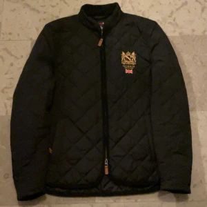 Svart jacka från Morris  - Snygg svart quiltad jacka från Morris med broderad logga och brittisk flagga på bröstet. Jackan har dragkedja framtill och långa ärmar med knappar vid ärmsluten. Perfekt för en stilren look. Skick 9/10 pris är förhandlingsbart 