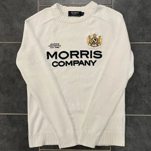 Vit tröja från Morris - Säljer en stilren vit tröja från Morris med broderad logotyp och text på framsidan. Tröjan är tillverkad i 100% bomull och har långa ärmar samt ribbade muddar vid hals, ärmslut och nederkant. Perfekt för en klassisk look.