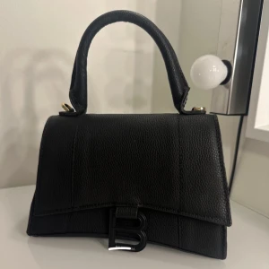 Svart handväska med axelrem - Snygg svart handväska i skinn med en stilren design och ett stort B som detalj framtill. Väskan har en praktisk avtagbar axelrem och ett robust handtag. Perfekt för att ge din outfit en elegant touch.