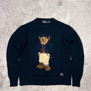 Intressekoll! - Säljer en mörkblå stickad tröja från Polo Ralph Lauren med en broderad björn på framsidan. Tröjan har långa ärmar och ribbstickade muddar vid ärmslut och nederkant. Perfekt för en stilren och avslappnad look. Kom med bud!