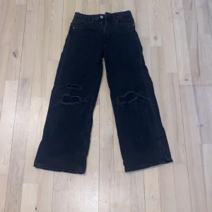 Svarta jeans med slitningar - Säljer ett par svarta jeans med coola slitningar på benen och en liten slits vid fållen. De har en klassisk femficksdesign och stängs med dragkedja och knapp. Perfekta för en avslappnad stil. För 9-10 åringar!!!