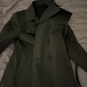 Svart trenchcoat från Tiger of Sweden - Snygg svart trenchcoat från Tiger of Sweden med klassisk design och knappar framtill. Perfekt för en stilren look. Fodrad insida för extra komfort.