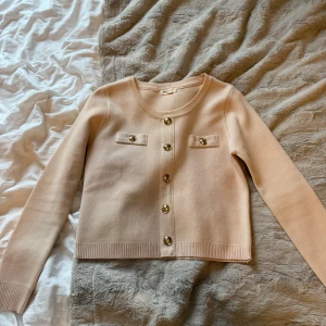 Beige kofta från Modocci - Säljer en stilren beige kofta från Modocci med dekorativa knappar i guld. Koftan har långa ärmar och ribbade kanter vid ärmslut och nederkant. Perfekt för en elegant look. Nypris 500kr, pris kan diskuteras