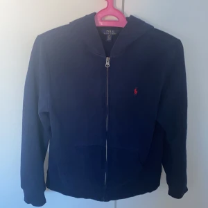 Mörkblå hoodie från Polo Ralph Lauren - Säljer en snygg mörkblå hoodie från Polo Ralph Lauren med dragkedja och det klassiska röda logotypen på bröstet. Tröjan har långa ärmar och en praktisk huva. Perfekt för en avslappnad stil. Jag är 167 cm och den sitter ganska tajt på mig den skulle passa någon runt 153-163 cm. Den har en liten defekt vid högra armen men de är inget man märker priset kan absolut diskuteras 