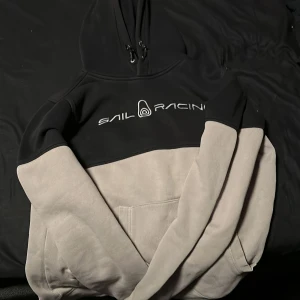 Svart och grå hoodie från Sail Racing - Säljer en snygg svart och grå hoodie från Sail Racing. Den har en stor ficka framtill och justerbar huva med dragsko. Perfekt för kyliga dagar och avslappnade stunder. 🖤