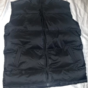 Svart dunväst med dragkedja från Weekday - Säljer en svart dunväst med dragkedja framtill. Västen har en enkel och stilren design med två fickor på sidorna. Perfekt för kyligare dagar när du vill hålla värmen utan att bära en jacka. Lite sminkfläckar längst kragen men annars som ny!