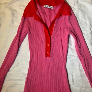 Rosa ribbad klänning från ADOORE - Snygg rosa ribbad klänning från ADOORE med  röd krage och knappar framtill. Klänningen har långa ärmar och en figurnära passform som ger en stilren look. Knappt använd 