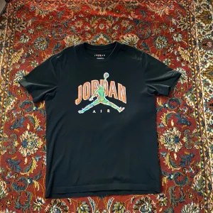 Svart Jordan t-shirt med grafiskt tryck - Säljer en svart Jordan t-shirt med ett färgglatt grafiskt tryck på framsidan. T-shirten har en klassisk rund hals och korta ärmar.