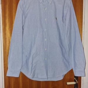 Blå skjorta från Ralph Lauren - Snygg blå skjorta från Ralph Lauren med klassisk design och knappar framtill. Perfekt för en stilren look. Den har långa ärmar och en liten broderad logga på bröstet.