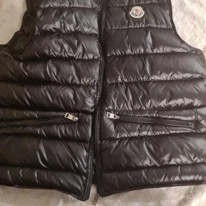 Svart dunväst från Moncler - Säljer min Moncler gui gillet svart. Förra ägaren gjorde tyvär sönder dragkedjan på mitten men alla fickor fungerar. Förra ägaren skrev också sitt namn och number i jackan på undersiddan av tagsen. Men det fungerar att scanna qr kodden. Namnet är också över struket så det är bara svart, kontakta för fler bilder.
