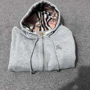 Grå hoodie från Burberry - Säljer en stilren grå hoodie från Burberry med dragkedja och klassiskt rutigt mönster i huvan. Tröjan har långa ärmar och ribbade muddar vid ärmslut och nederkant. Perfekt för en avslappnad och trendig look. .Sitter som s men är m. Hör av dig vid några frågor eller funderingar 