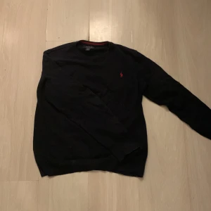 Marinblå tröja från Ralph Lauren - Säljer en Marinblå tröja från Ralph Lauren i mjuk pima bomull. Tröjan har en röd broderad logotyp på bröstet och ribbade kanter. Perfekt för en stilren look. Tröjan är XL i barn storlek så sitter som Xs