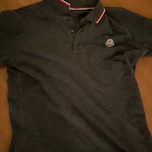 Grå pikétröja från Moncler - Säljer en stilren grå pikétröja från Moncler med klassisk krage och knappar. Tröjan har en broderad logotyp på bröstet och röd-vit randig detalj på kragen. Perfekt för en avslappnad men ändå elegant look. Du kan bekräfta autenticitet via QR kod på sista bilden .