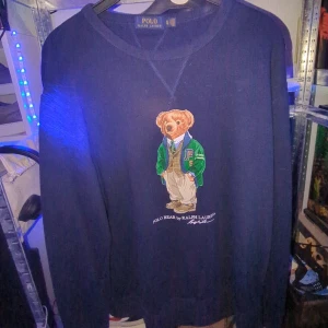 Ralph Lauren polo bear  - Säljer en mörkblå tröja från Ralph Lauren med ett ikoniskt björntryck på framsidan. Tröjan är tillverkad i en bomulls- och polyesterblandning och har långa ärmar. Perfekt för en stilren och avslappnad look. Inga skador som jag kan se. Xl i barn sitter som typ M-L skulle jag säga om ni tvättar den först lite ut töjd men aja. 
