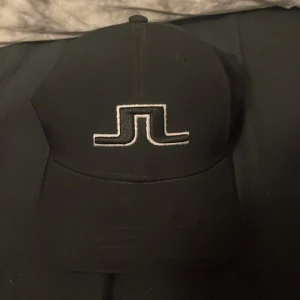 J.Lindberg nytt skick, aldrig använd - Snygg svart keps med en stilren broderad logga framtill. Perfekt för en casual look och passar till många olika outfits. Kepsen har en klassisk design med böjd skärm och justerbar passform.