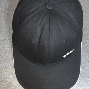 Svart keps från Boss - Snygg svart keps från Boss med vit logga på framsidan. Kepsen har en klassisk design med rundad skärm och ventilationshål för extra komfort. Perfekt för en stilren look.