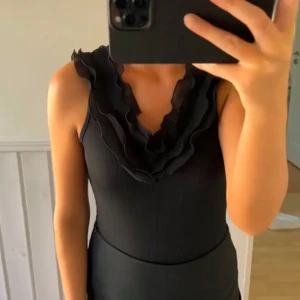 Svart ärmlös body med volangdetaljer - Snygg svart ärmlös body med volangdetaljer runt halsringningen och en öppen rygg. Perfekt för en stilren look.