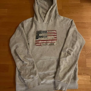 Ralph Lauren hoodie - Säljer en grå hoodie från Ralph Lauren med en vintageinspirerad amerikansk flagga på bröstet. Tröjan har en klassisk huva och långa ärmar, perfekt för en avslappnad stil. Hör av dig vid intresse!