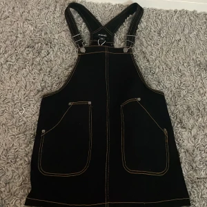 Svart klänning med hängslen från Monki - Snygg svart klänning från Monki med justerbara hängslen och kontrasterande sömmar. Den har två stora fickor framtill och är perfekt för en avslappnad stil. Klänningen är i en bekväm passform och passar bra till olika tillfällen.