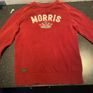 Röd tröja från Morris - Snygg röd tröja från Morris med text och brittisk flagga på framsidan. Tröjan har långa ärmar och ribbade muddar vid ärmslut och nederkant. Perfekt för en stilren look.