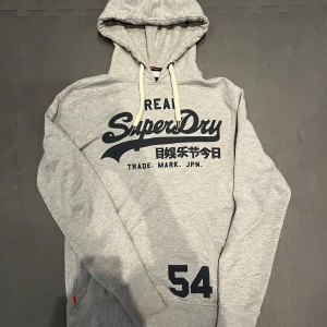 Superdry hoodie  - Säljer en riktigt fin hoodie från märket Superdry, den är i fint skick och den ända defekten är på vänstra huvsnöret där lite nopper och lösa trådar förekommer, bilder kan skickas privat. Varm och skön och perfekt året om, säljs inte längre och mycket svår att få tag på i såhär fint skick. Skriv vid minsta fundering eller om du har frågor!🤝🤝 pris kan självklart diskuteras vid smidig affär!