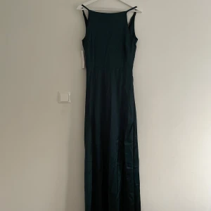 Mörkgrön långklänning i satin - Elegant mörkgrön långklänning i satin från Bubbleroom. Klänningen har smala axelband och en stilren design som passar perfekt för speciella tillfällen. Den är tillverkad i ett mjukt och glansigt material som ger en lyxig känsla.