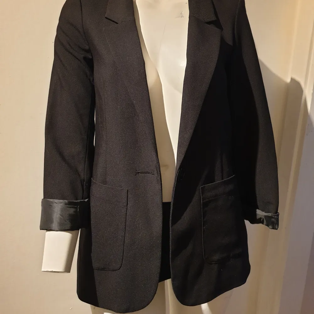 Elegant svart kavaj med klassisk design och knappar framtill. Kavajen har långa ärmar och fickor på sidorna, vilket ger en stilren look. Perfekt för en formell eller professionell stil.. Puvut & Bleiserit.