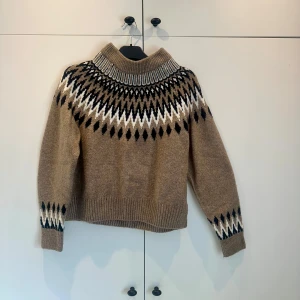 Beige stickad tröja med mönster - Säljer en supermysig beige stickad tröja från H&M i stel XS, ett snyggt mönster i svart och vitt runt halsen och ärmsluten. Perfekt för kalla dagar och ger en härlig vinterkänsla. Tröjan har långa ärmar och en ribbad kant vid ärmslut och nederkant. 