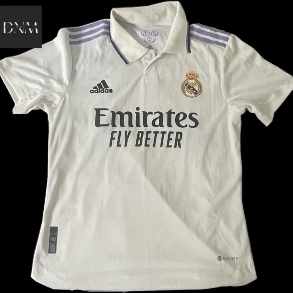 Säljer en vit Real Madrid fotbollströja från Adidas. Tröjan har korta ärmar och är i nyskick. Den har klubbens emblem på bröstet och sponsorloggan 'Emirates Fly Better' tryckt på framsidan. På baksidan står det 'VINI JR.' med nummer 20. . T-shirts.