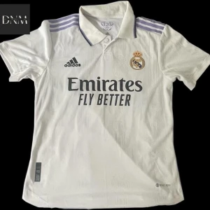 Vit Real Madrid fotbollströja - Säljer en vit Real Madrid fotbollströja från Adidas. Tröjan har korta ärmar och är i nyskick. Den har klubbens emblem på bröstet och sponsorloggan 'Emirates Fly Better' tryckt på framsidan. På baksidan står det 'VINI JR.' med nummer 20. 