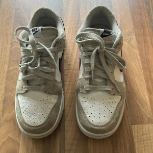 Nike Dunk i mocka och skinn - Säljer ett par Nike Dunk sneakers i en snygg kombination av beige mocka och vit skinn. Skorna har en klassisk design med svarta och turkosa detaljer, inklusive den ikoniska Nike-loggan på sidan. De är i bra skick med lite slitage på sulorna. Perfekta för både vardag och fest!