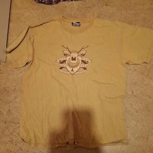 Beige t-shirt med tryck - Säljer en beige t-shirt med ett coolt tryck på framsidan. Den är kortärmad och perfekt för en avslappnad stil. Passar bra till jeans eller shorts för en casual look. Perfekt för sommardagar eller som en del av en lager-på-lager outfit.