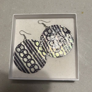 Runda örhängen - Snygga runda örhängen med ett glansigt mönster i svart och silver. De har en cool design med cirklar och ränder som verkligen sticker ut. Perfekta för att ge din outfit en extra touch av glamour. Passar både till fest och vardag!