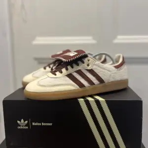 Säljer ett par Adidas Wales Bonner sneakers i mycket bra skick. Skorna är i en snygg beige färg med mörkbruna detaljer och klassiska tre ränder på sidan. De har en gummisula och snörning framtill. Perfekta för både vardag och fest!