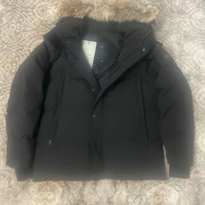 Svart dunjacka från Canada Goose - Säljer en svart dunjacka från Canada Goose i mycket bra skick. Jackan har en avtagbar huva med fuskpäls och långa ärmar. Perfekt för kalla vinterdagar med sin varma och isolerande design. Den har både dragkedja och knappar för extra skydd mot kyla. Storleken är M men har flera i olika storlekar 