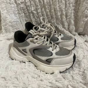 Grå och vita sneakers - Säljer ett par grå och vita sneakers i mycket bra skick. De har en chunky sula och snyggt meshmaterial som ger en sportig look. Perfekta för både vardag och träning. Skorna har snörning och är riktigt bekväma att gå i. Passar perfekt till jeans eller träningskläder! Liknar New balance, endast använda 2 gånger, nypris 449kr