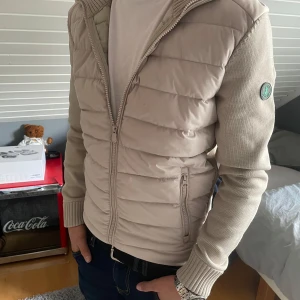 Beige jacka från Save the Duck - Säljer en snygg beige jacka från Save the Duck i mycket bra skick. Jackan har en quiltad framsida med dragkedja och ribbstickade ärmar och krage. Perfekt för höst och vår! Använd Max 4 gånger!!!! Helt ny. Passar perfekt för folk som är 165 -175 
