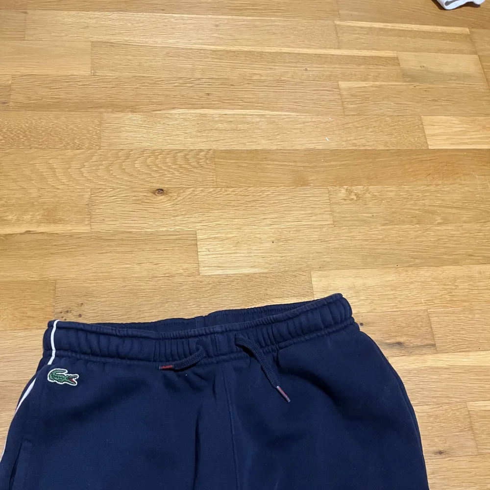 Säljer en snygg vit träningsjacka från Lacoste med marinblå och röd rand över bröstet. Jackan har dragkedja och långa ärmar med ribbade muddar. Perfekt för sportiga aktiviteter eller en casual look. Passar både vår och höst!. Tröjor & Koftor.