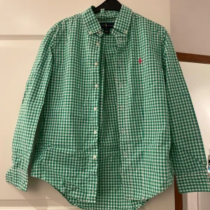 Grön och vit rutig skjorta från Ralph Lauren - Säljer en snygg grön och vit rutig skjorta från Ralph Lauren i mycket bra skick. Skjortan har långa ärmar och knappar framtill. Den är perfekt för både vardag och festliga tillfällen. Broderad logga i rosa på bröstet ger en extra touch av stil. Passar perfekt för vår och sommar!