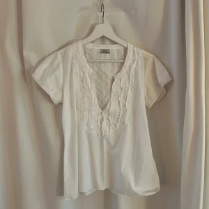 Vit blus från Hunkydory - Säljer en söt vit blus från Hunkydory i bra skick. Den har korta ärmar och fina volangdetaljer framtill. Perfekt för vår och sommar, och passar både till vardags och fest. Blusen är i ett lätt och luftigt material, vilket gör den bekväm att bära.