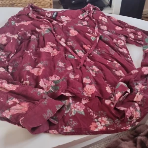Vinröd blommig blus från H&M Divided - Säljer en superfin vinröd blus från H&M Divided i storlek 34. Den har ett vackert blommigt mönster i rosa och grönt. Blusen har långa ärmar med knappar vid ärmsluten och en avslappnad passform. Perfekt för både vår och höst!
